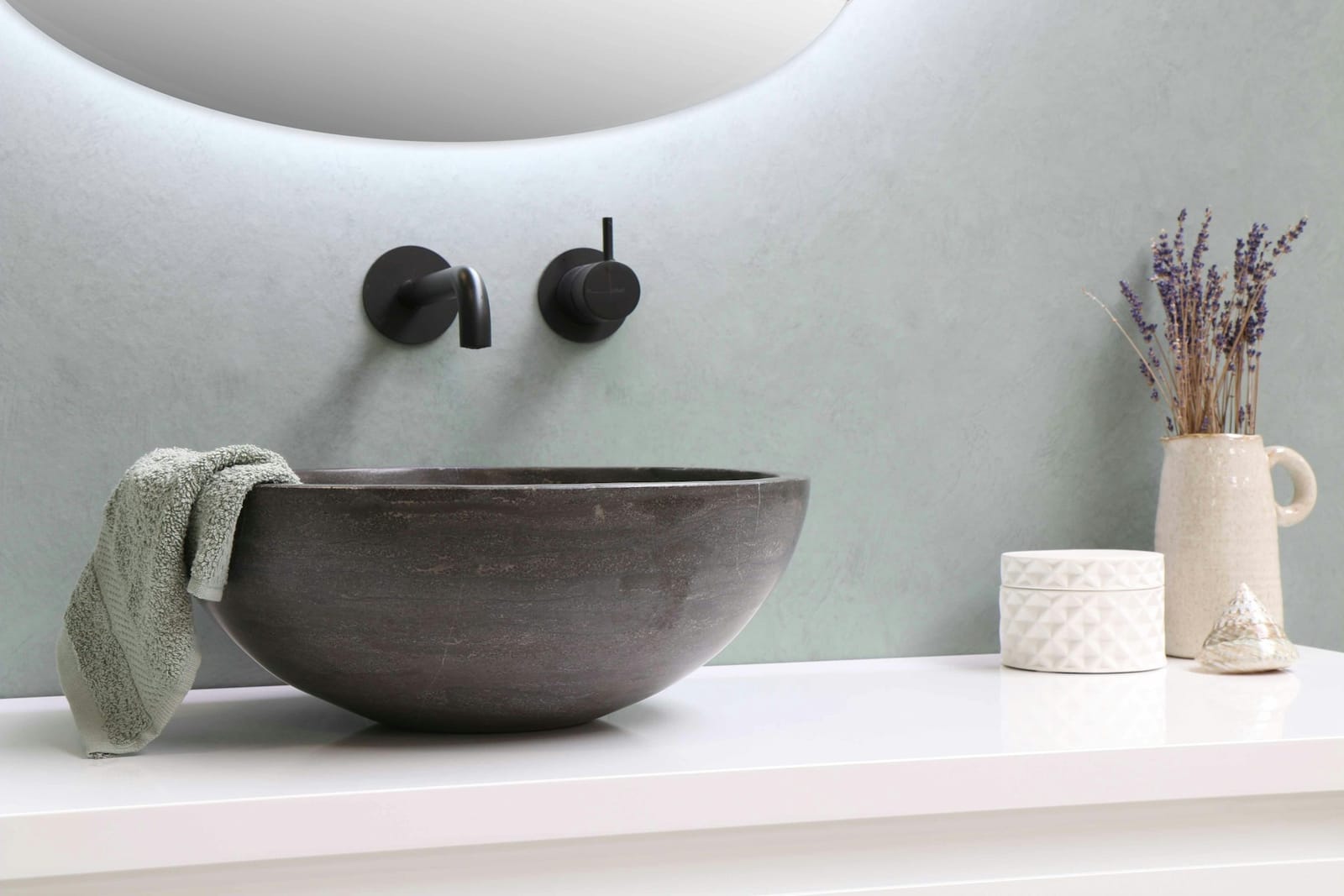 Bagno con lavabo a bacinella in pietra scura, specchio retroilluminato, rubinetteria nera e asciugamano verde salvia